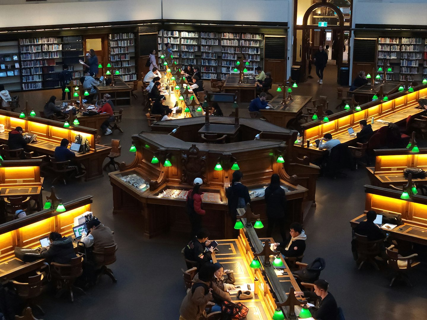Students and patrons study at green-lamp lit desks in a grand library.